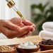 Close-up of a massage therapist catching a golden drop of CBD oil from an amber dropper bottle, with bowls of jojoba seeds, coconut pieces, and grapeseeds on a wooden tray and soft towels and greenery blurred in the background.