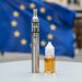 A modern vaping device and capped e-liquid bottle on a clean tabletop with a softly blurred EU flag in the background.